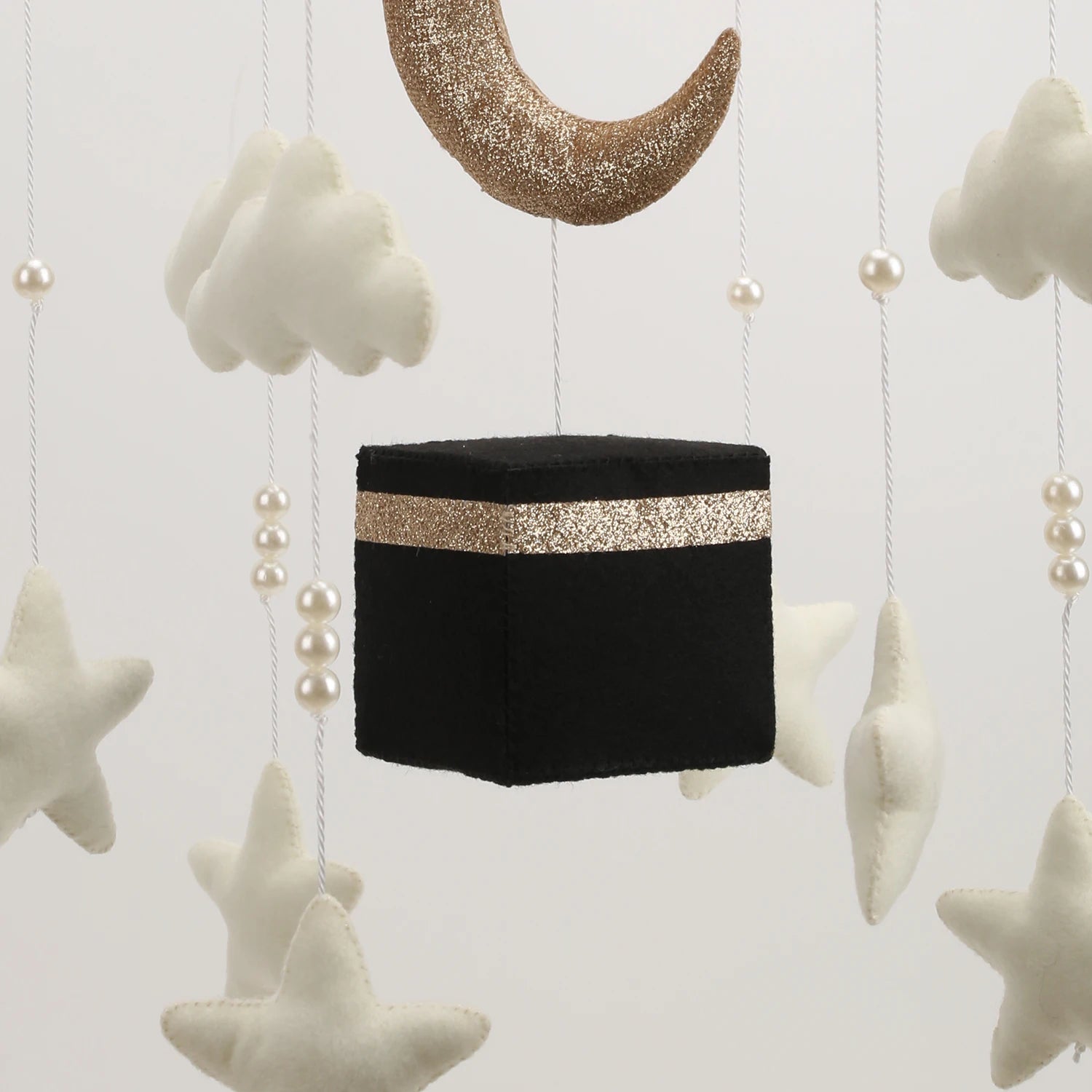 Decorative hanging item resembling a Kaba with gold band, surrounded by stars and clouds on a light background.